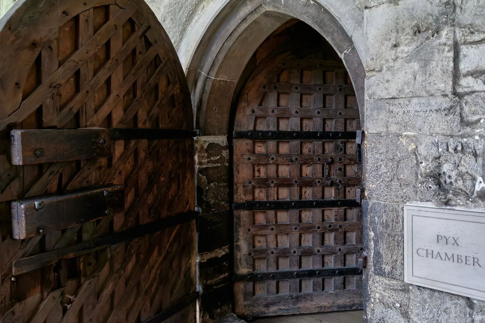 An ancient studded oak door within a pointed Gothic arch beside a stone wall marked Pyx Chamber at Westminster Abbey