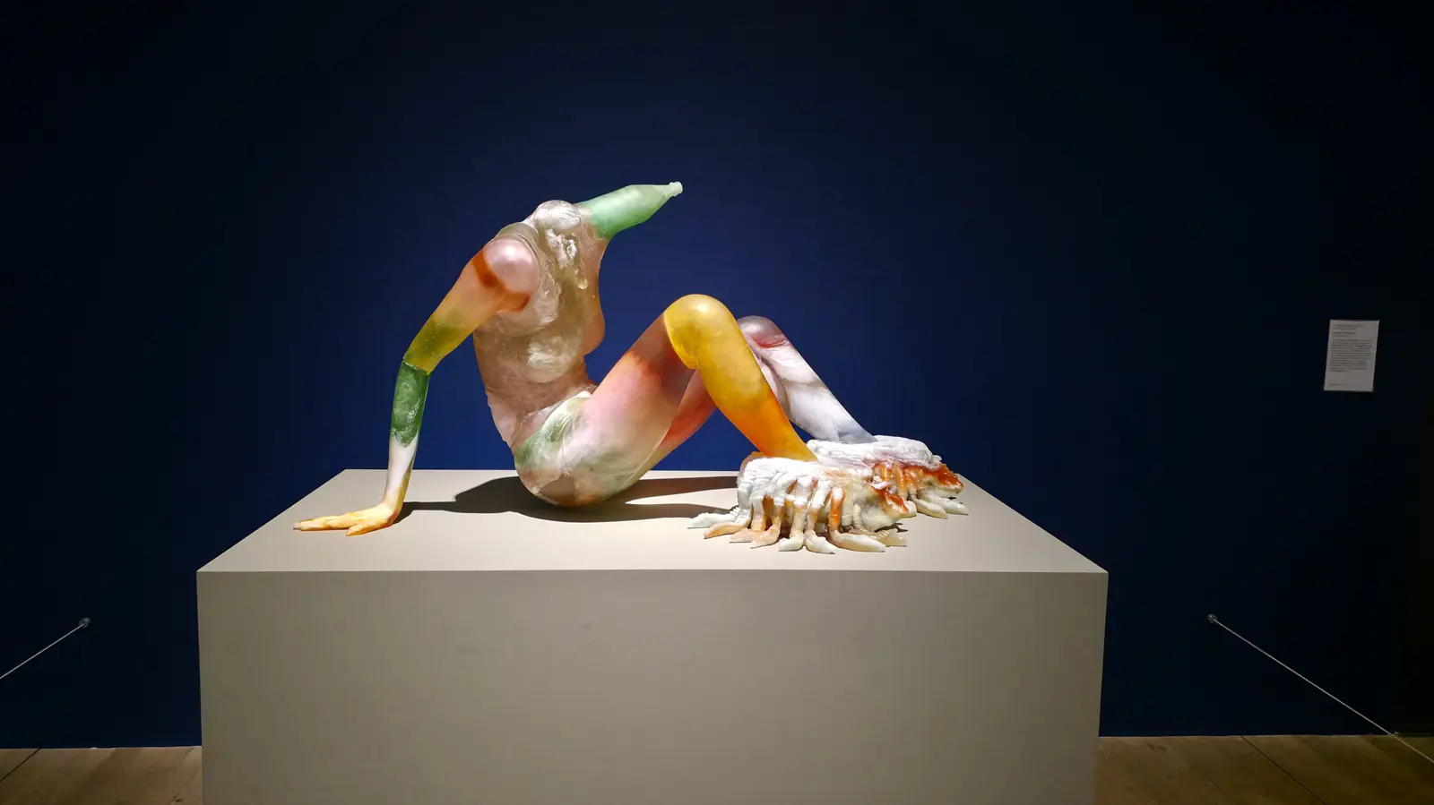 Andra Ursuța’s Predators ’R Us 2020, a reclining figure in multicoloured cast glass, lit against a deep blue wall at Tate Modern