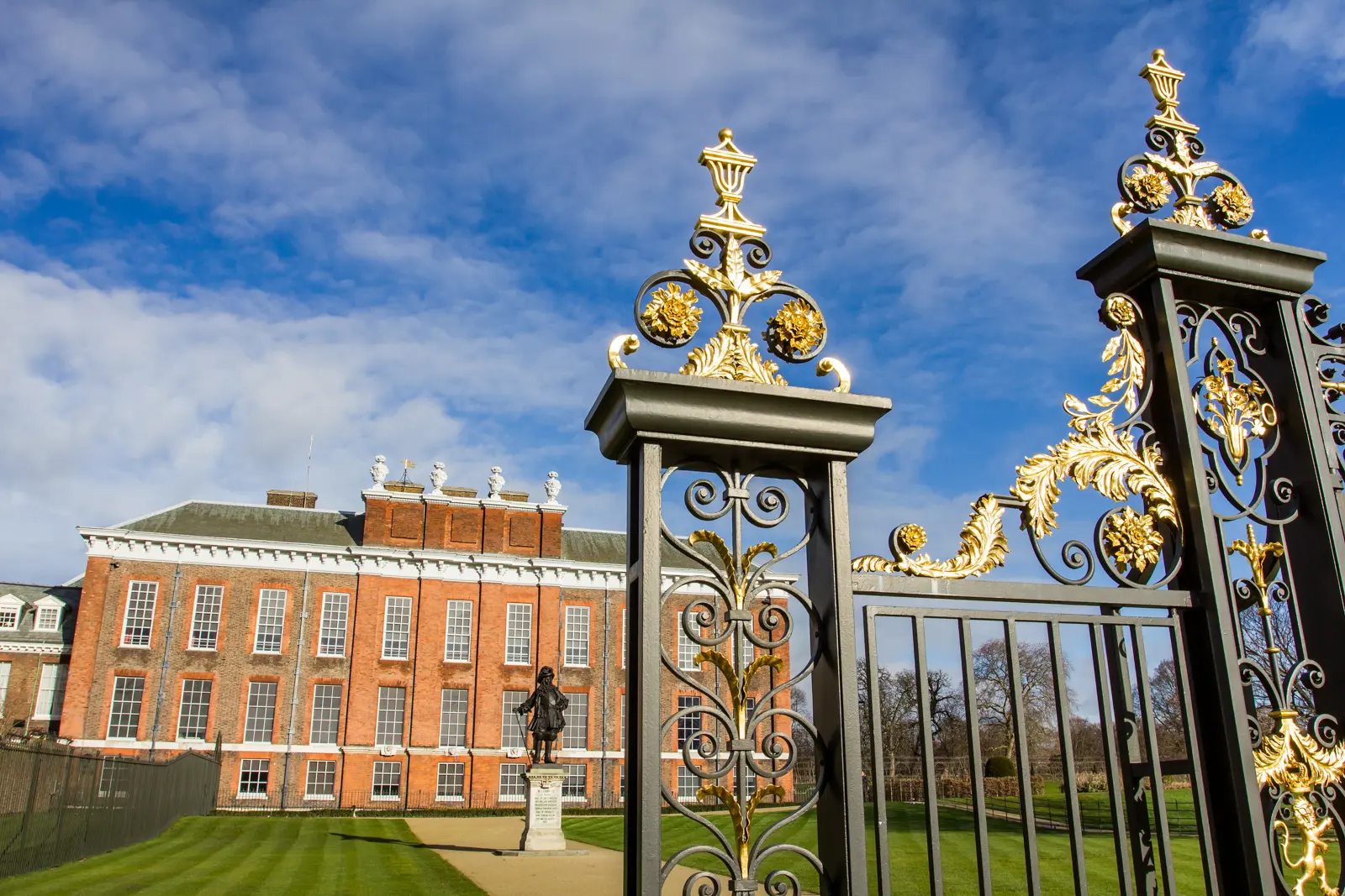 Kensington Palace behind its gilded wrought-iron gates, with the bronze statue of William III on the lawn, Kensington Gardens, London.