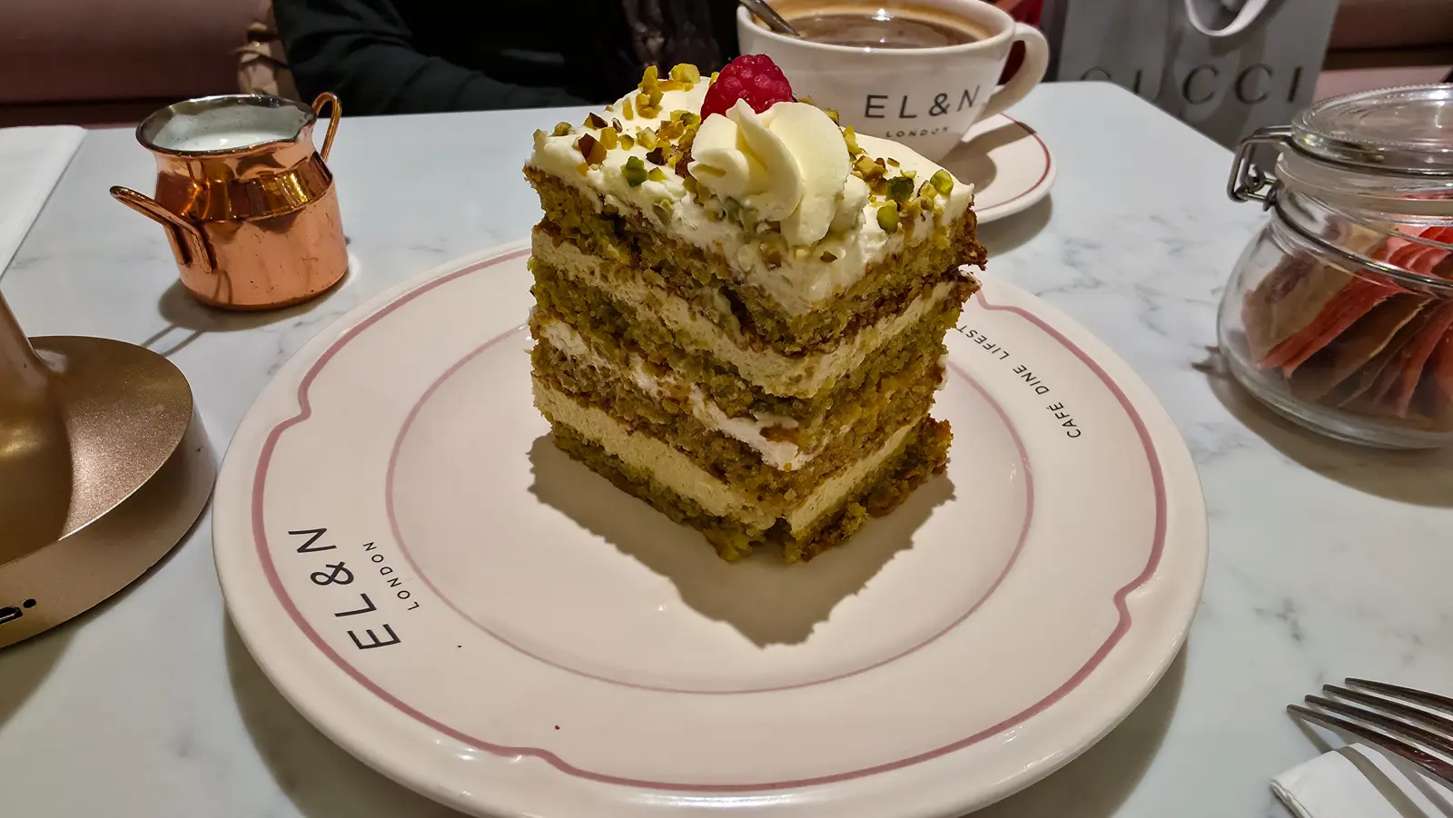 A slice of pistachio layer cake topped with cream and a raspberry, on a branded EL&N London plate in Knightsbridge