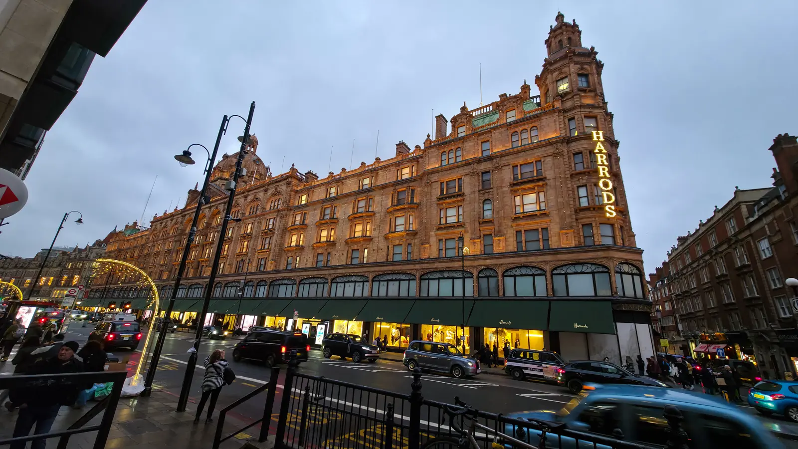 The terracotta facade of Harrods in Knightsbridge, London, lit up at dusk with taxis and traffic on Brompton Road