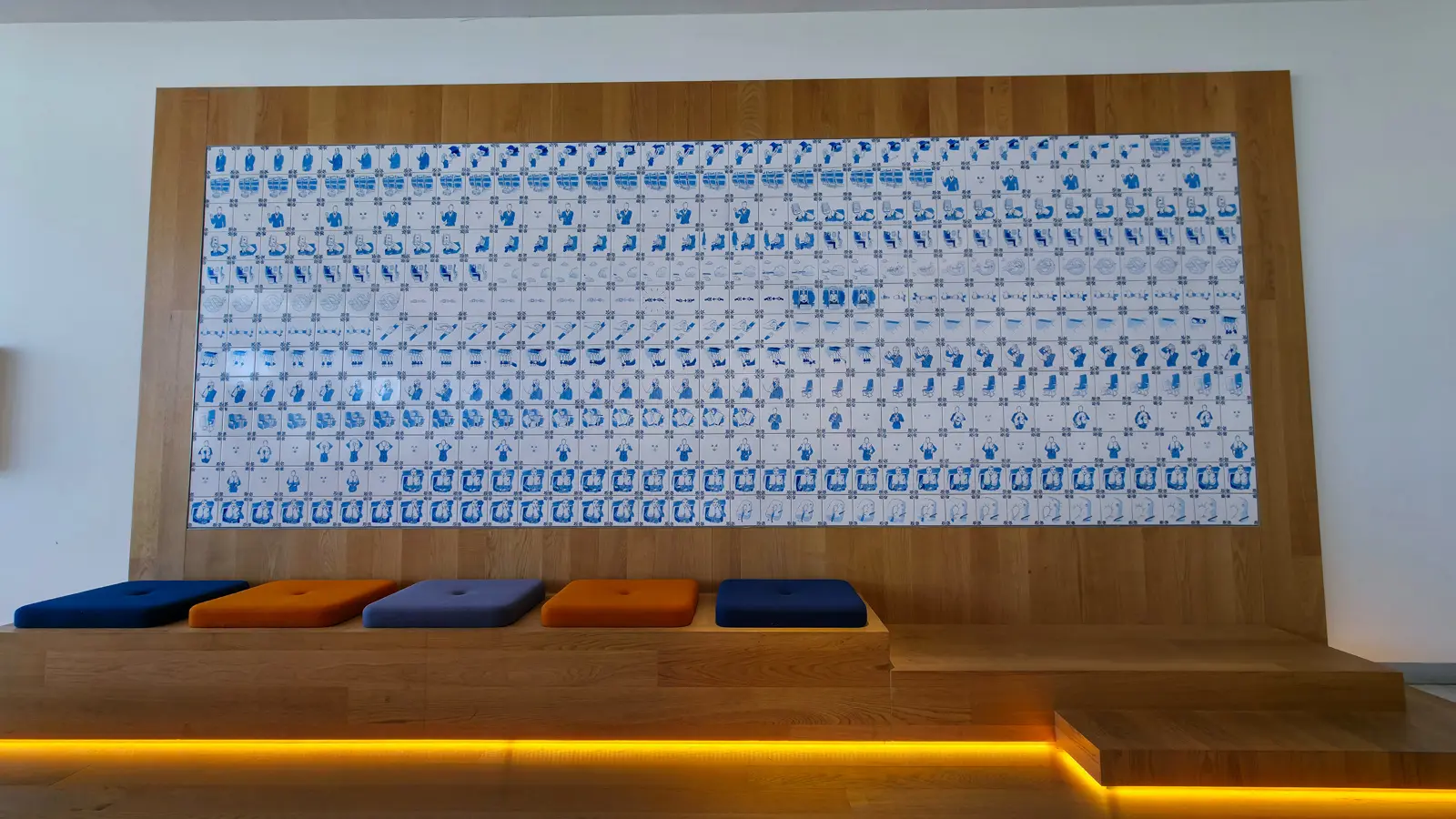 A large panel of blue and white Delft tiles showing safety-instruction figures, above a wooden bench with blue and orange cushions, Crown Lounge 52, Schiphol