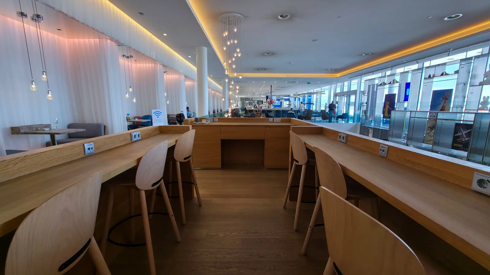 A long wooden work counter with stools, power sockets and pendant lights, beside vintage KLM posters, Crown Lounge 52, Schiphol