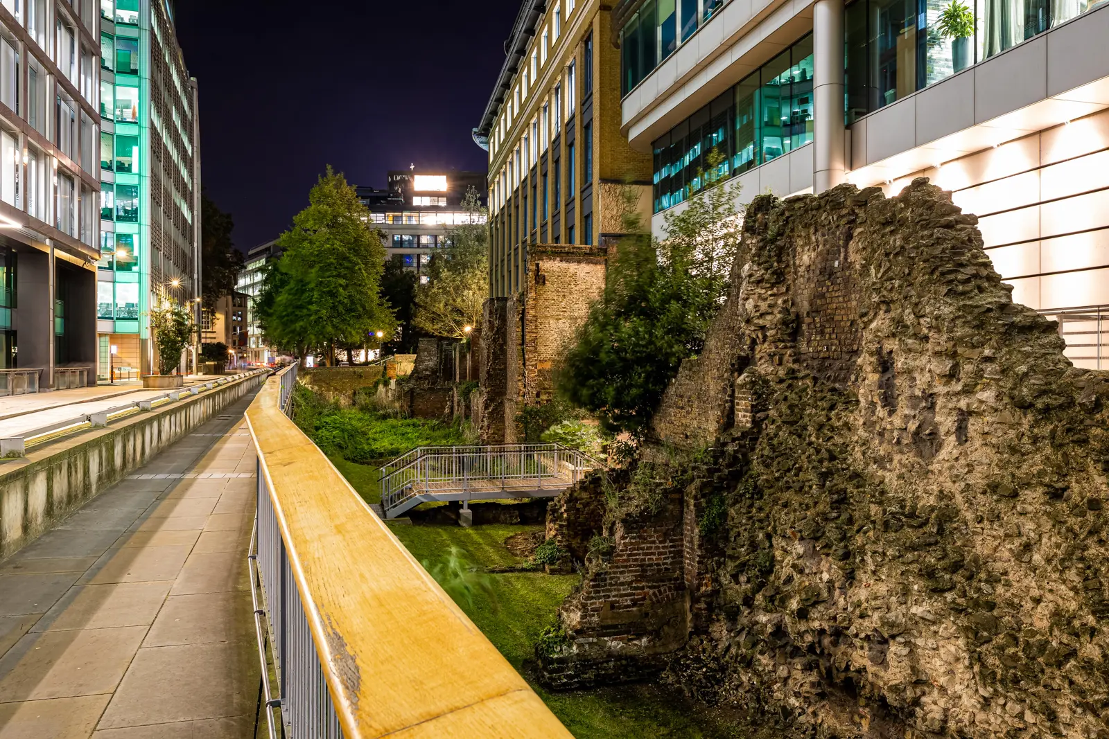 A floodlit section of the ancient London Wall at night, running between modern glass office buildings in the City