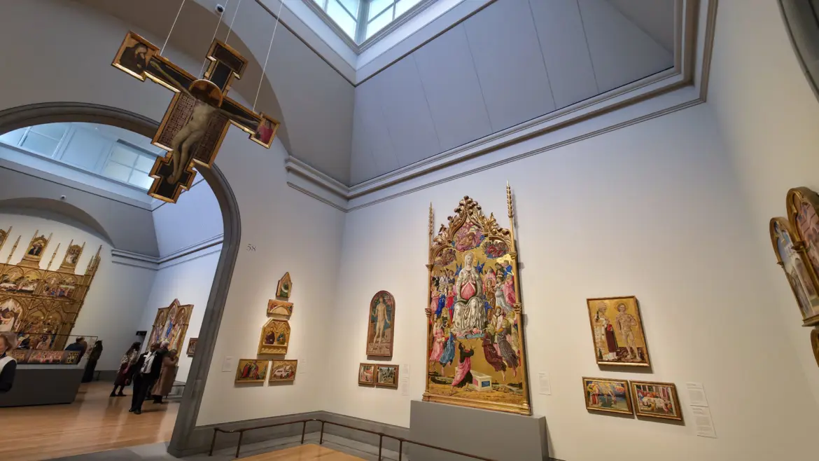 A tall gold-ground gallery at the National Gallery, London, with a painted crucifix suspended from the ceiling and altarpieces lining the walls.