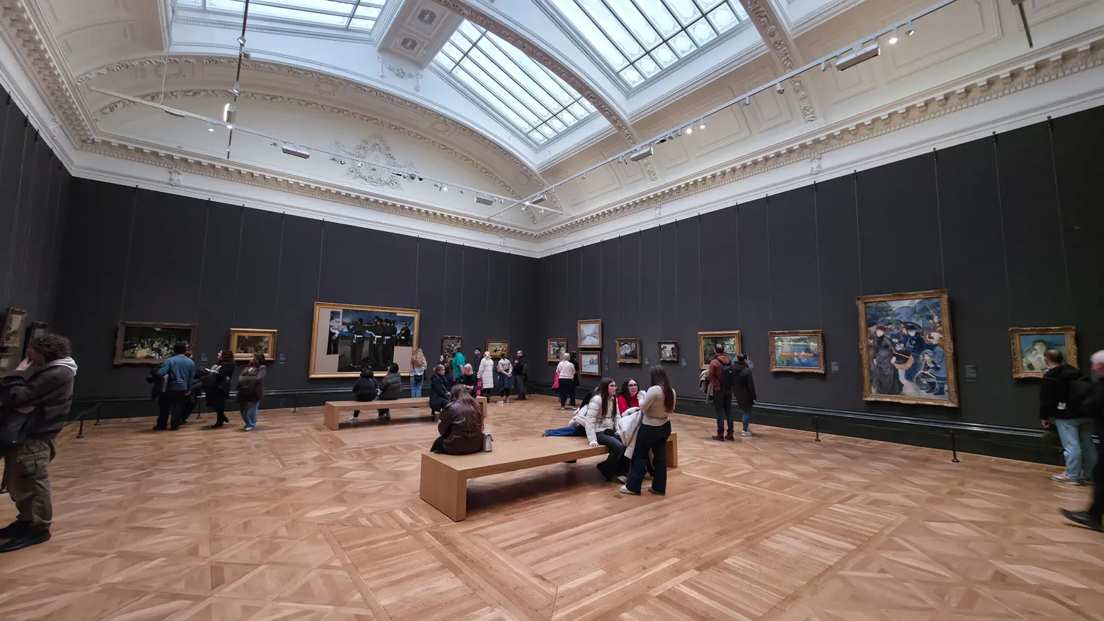 Gilt-framed Impressionist paintings on grey walls under a bright skylight in a gallery at the National Gallery, London
