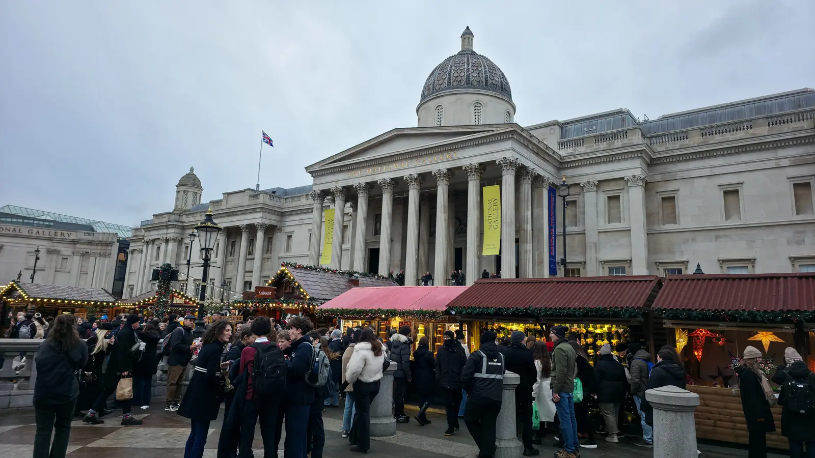The National Gallery above Trafalgar Square in London, with wooden Christmas market chalets and crowds in front of the portico