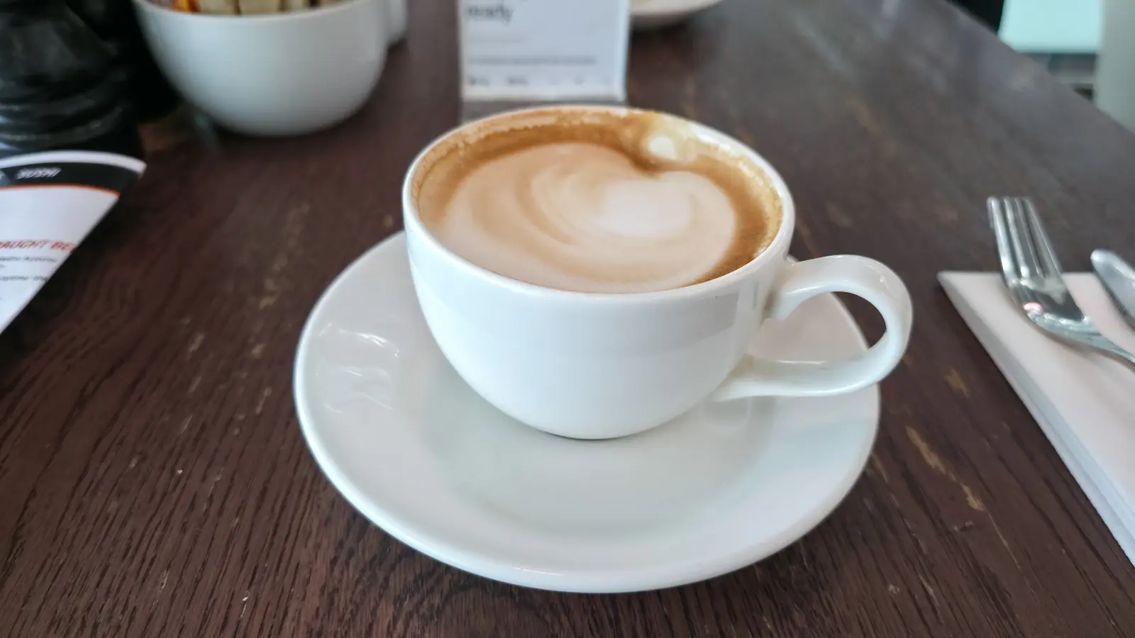 A white cup of coffee with a swirl of steamed milk on the surface, on a dark wood table at Gordon Ramsay Plane Food Market, Heathrow Terminal 5