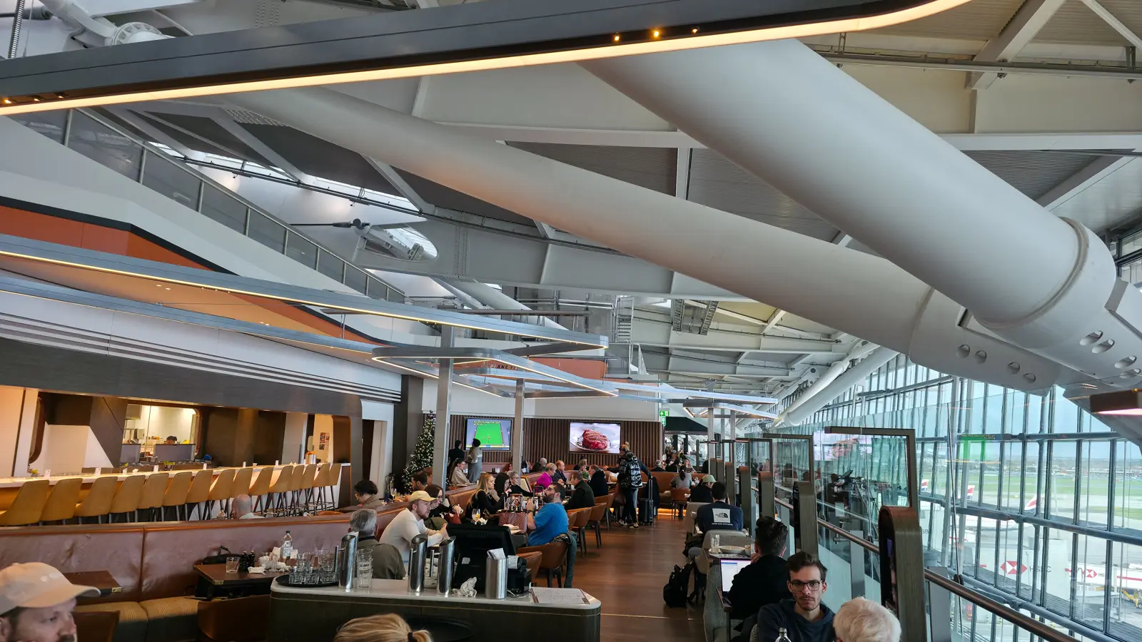 The dining room at Gordon Ramsay Plane Food Market, leather banquettes under Terminal 5's curved roof, planes beyond the windows