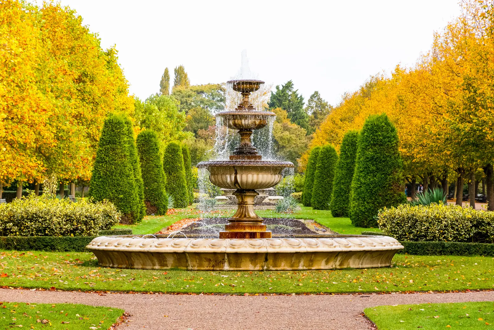 A tiered stone fountain running in the Avenue Gardens, framed by clipped conifers and autumn trees