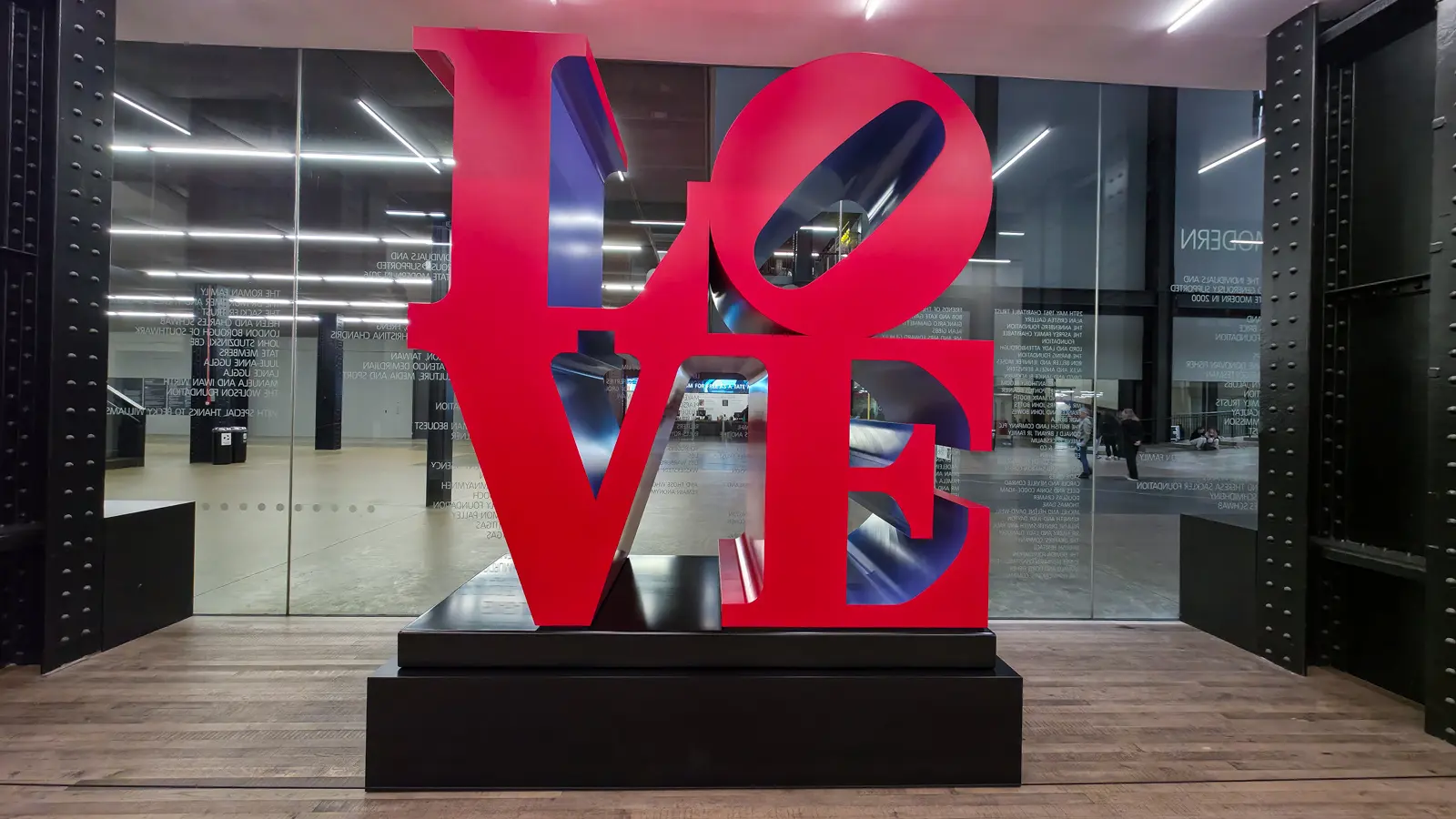 Robert Indiana’s LOVE sculpture in red with blue inner faces, on a black plinth beside a glass wall at Tate Modern