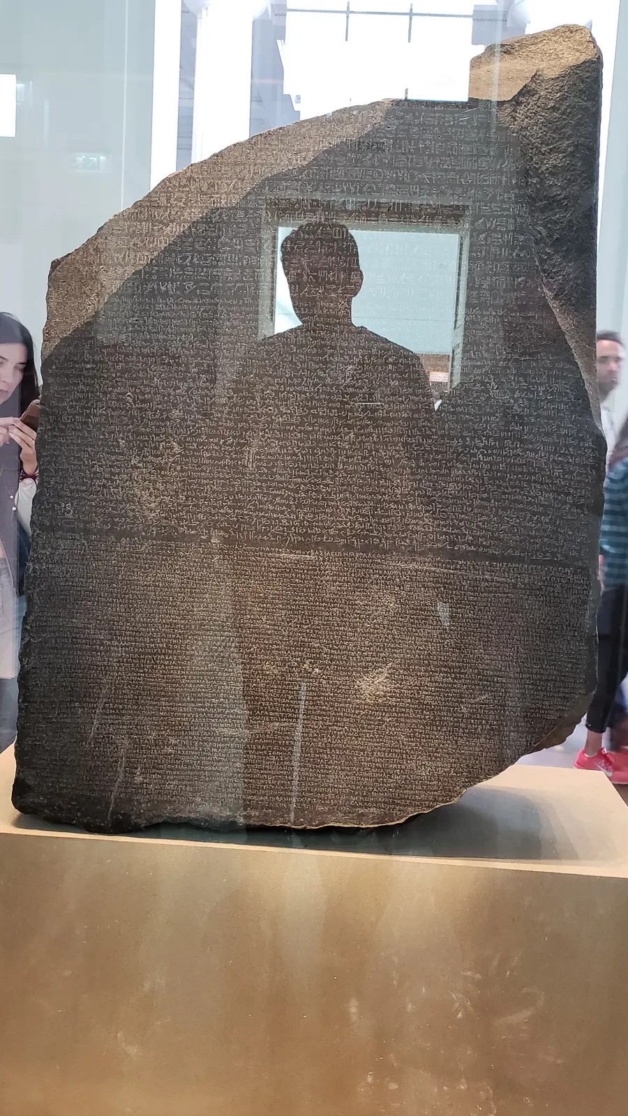 The full face of the Rosetta Stone with its three bands of inscription, a visitor’s silhouette reflected across the centre, British Museum
