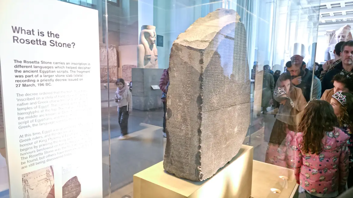The Rosetta Stone in its glass case in Room 4 of the British Museum, with visitors reflected across the surface