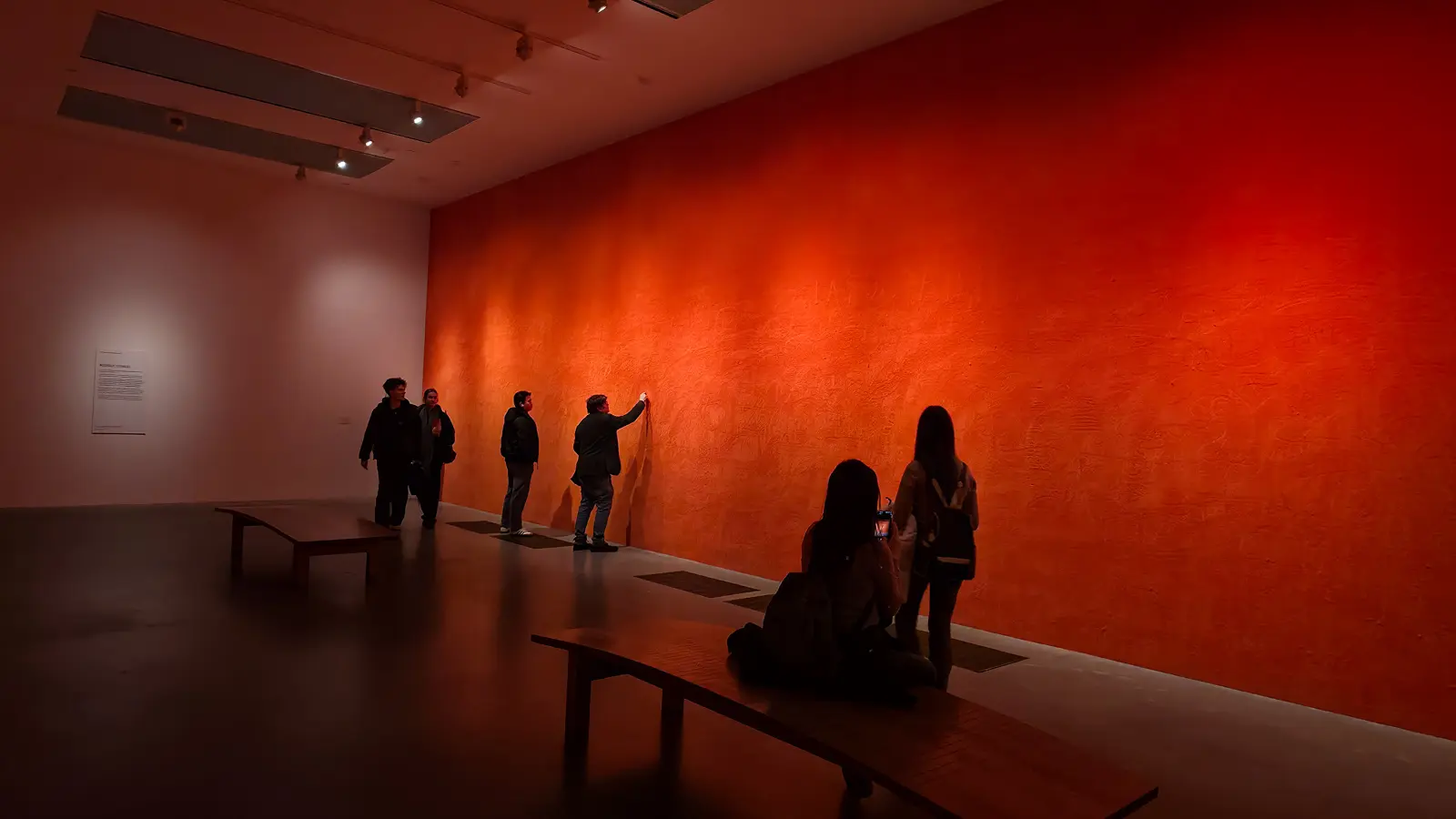 Visitors touching Rudolf Stingel’s orange carpeted wall work Untitled 1993 in a dimly lit gallery at Tate Modern, London