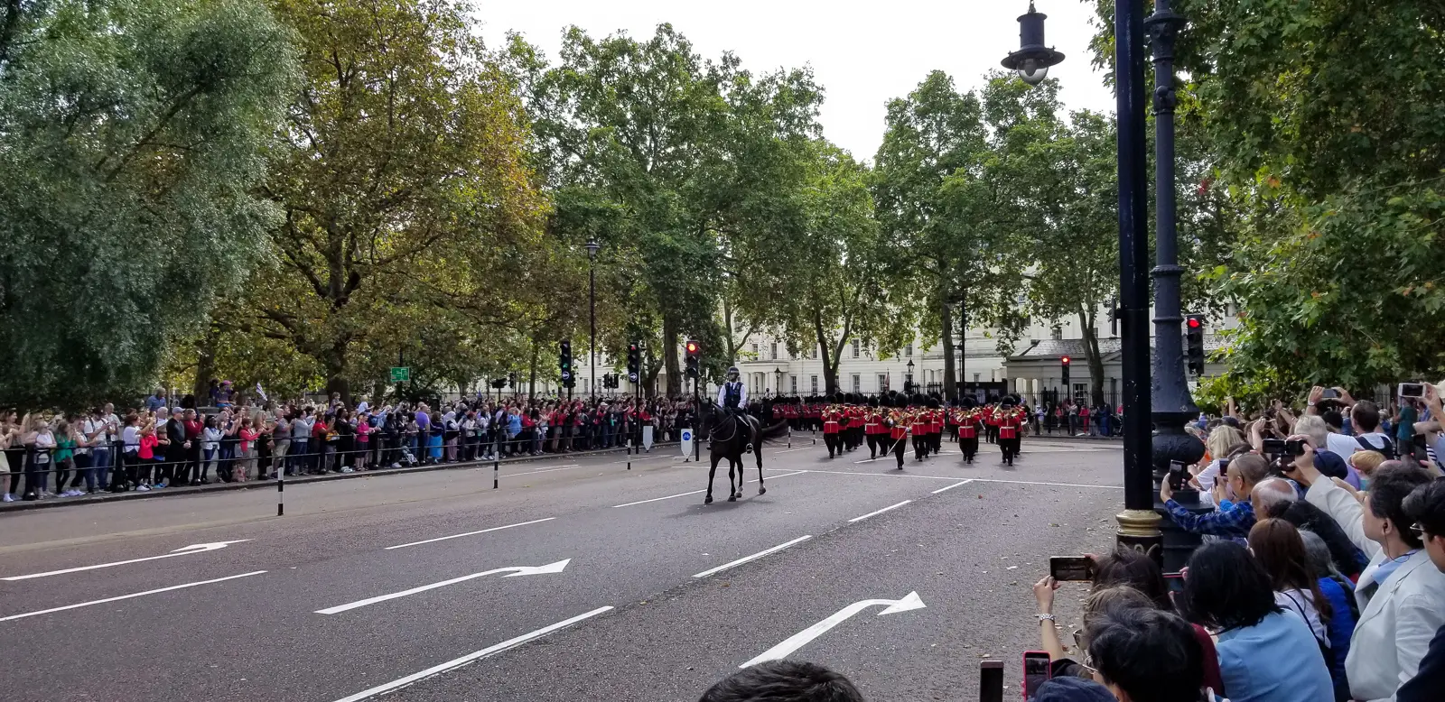 A military band in scarlet and bearskins marching past crowds with a mounted police escort during Changing the Guard, London.