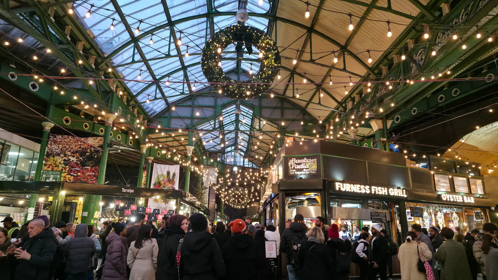 Borough Market under its glass roof at Christmas, with festoon lights, a giant wreath and food stalls including an oyster bar