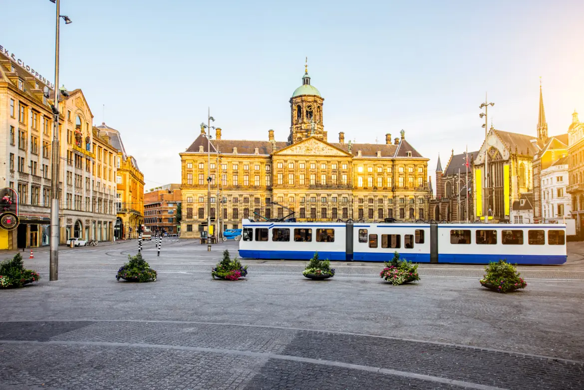 The Royal Palace Amsterdam in early morning light on Dam Square, with a tram passing and the Nieuwe Kerk to the right