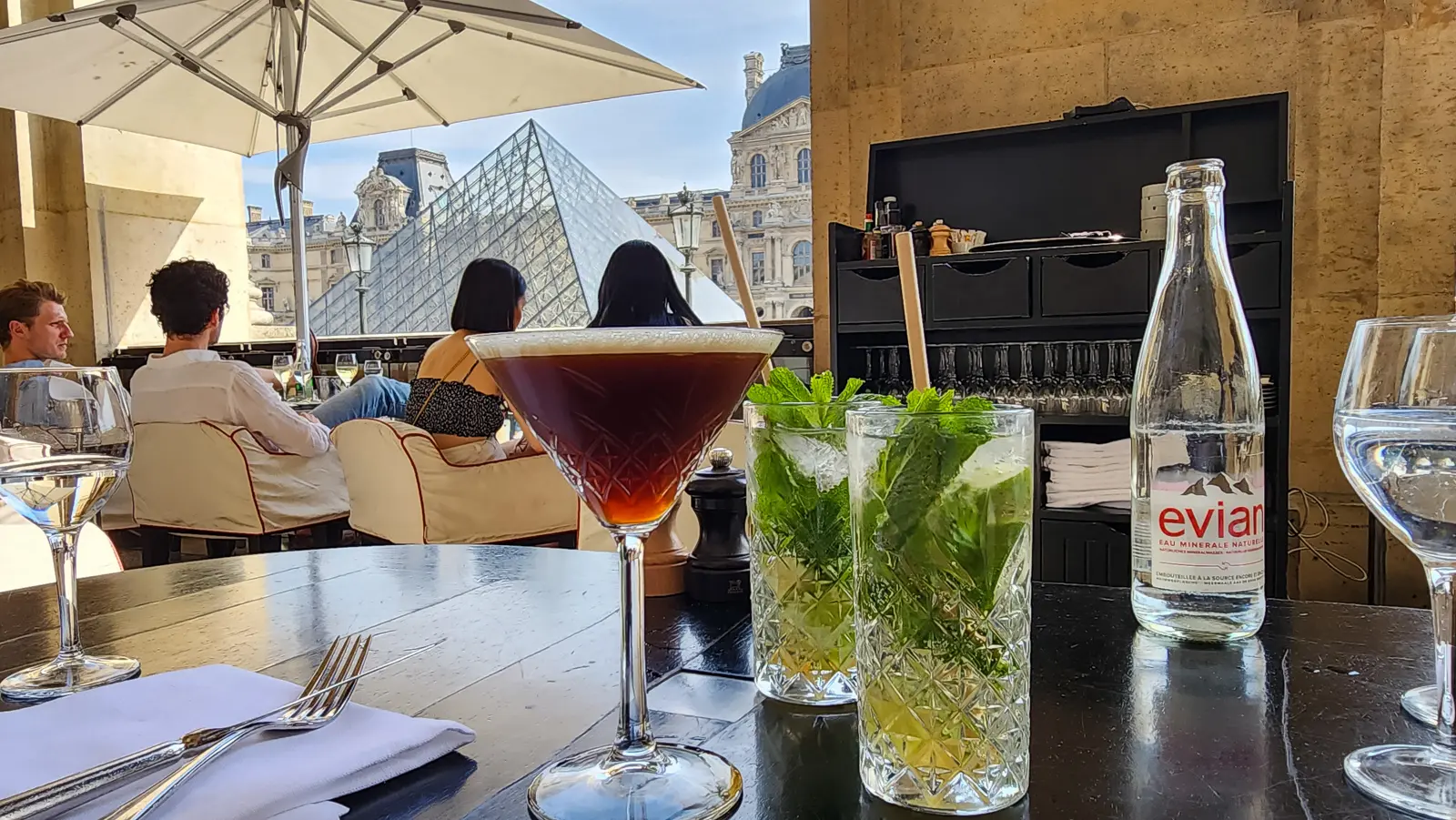 Drinks on a table at Cafe Marly under the arcade, with the glass pyramid and the Louvre’s wings visible past a white parasol