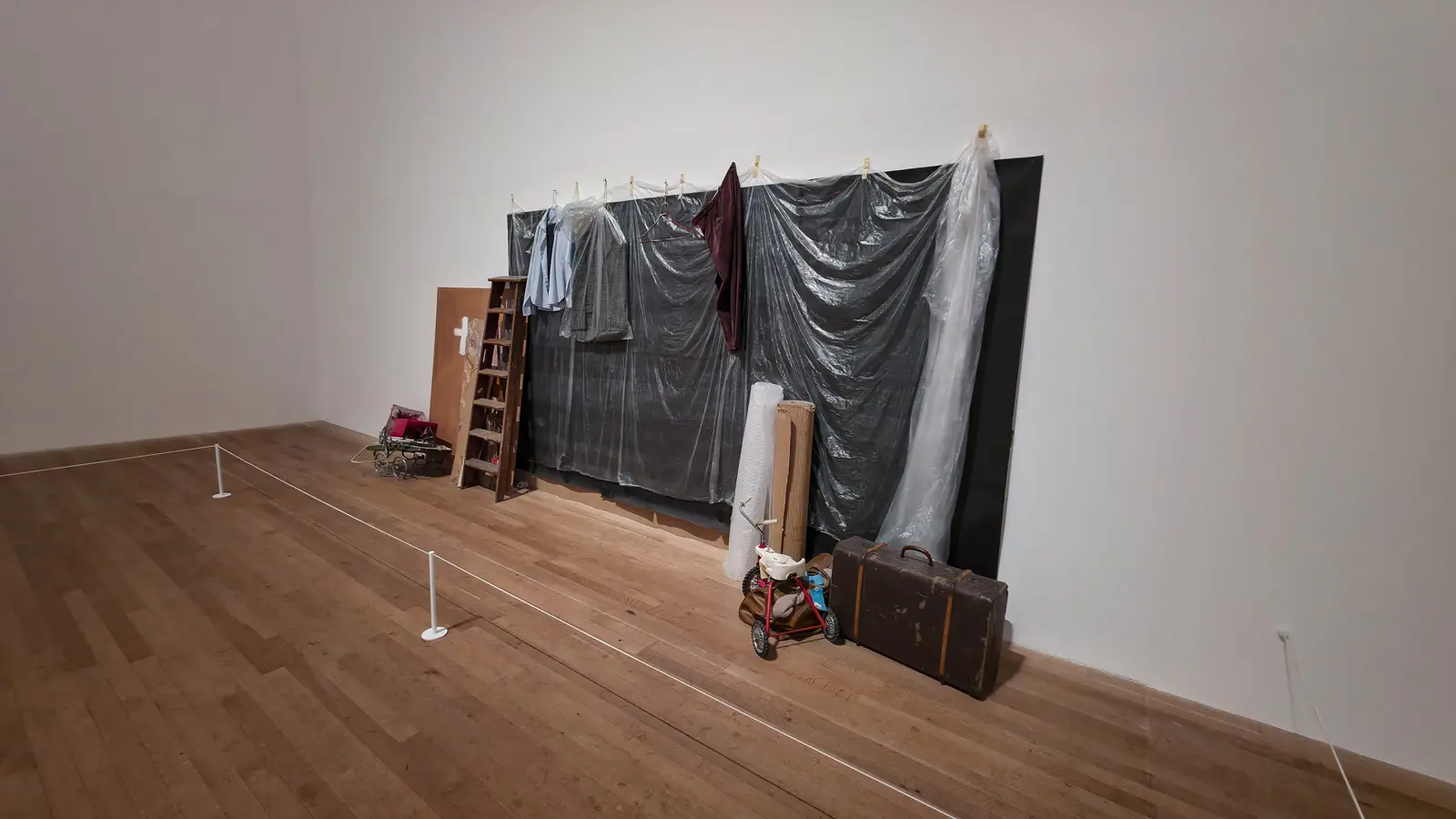 Vlassis Caniaris’s Possible Background at Tate Modern: plastic sheeting, hung clothing, a suitcase and a child’s tricycle