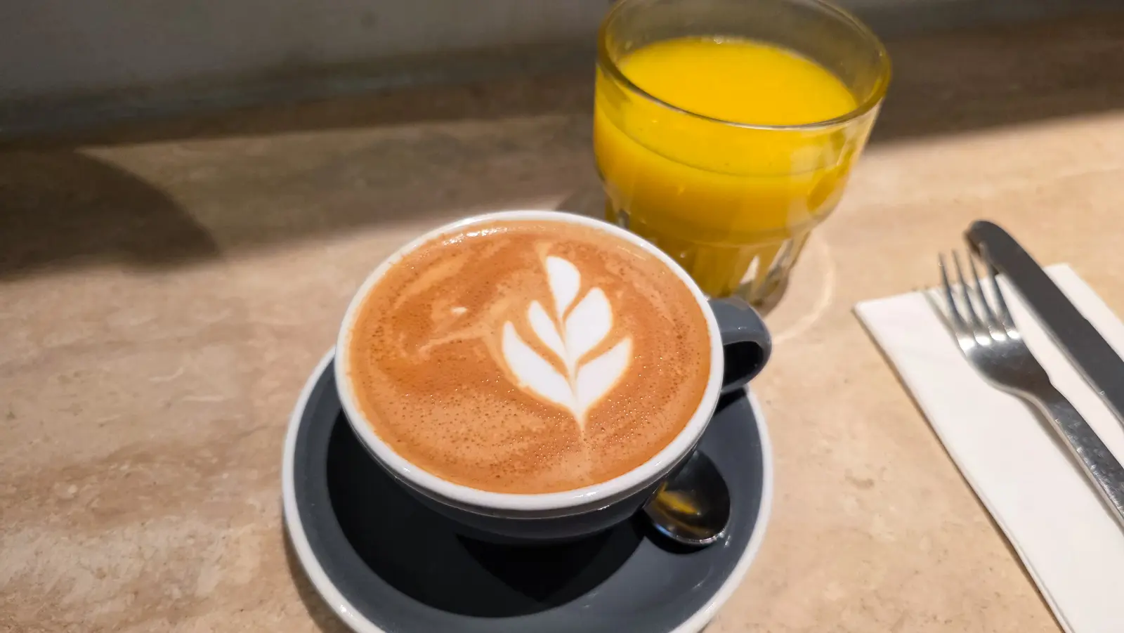 A cappuccino with a leaf poured into the foam, in a grey cup on a grey saucer, beside a glass of orange juice on a stone table