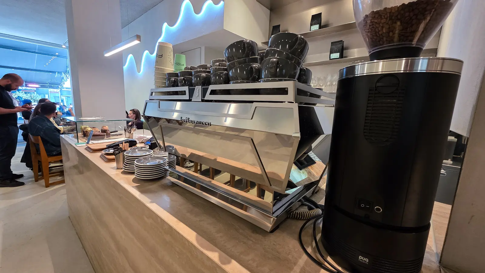 The counter at Flow with stacked grey cups on the espresso machine, saucers, a glass cake case and coffee bags on the shelves behind