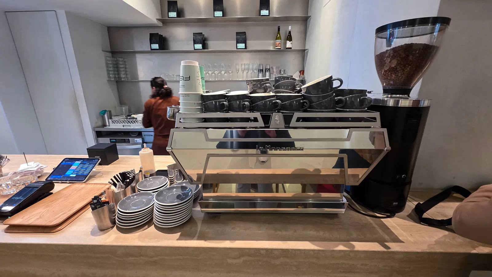 Flow’s espresso machine seen head on, with cups stacked above it, a grinder beside it, and shelves of coffee bags and wine bottles behind