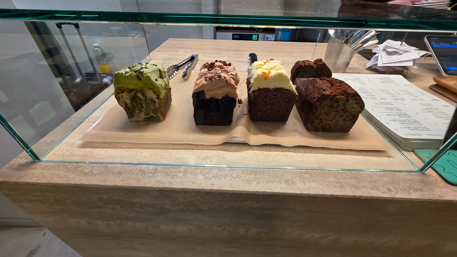 Four iced loaf cakes in a glass case on the counter at Flow, cut to show the section, on a sheet of baking paper