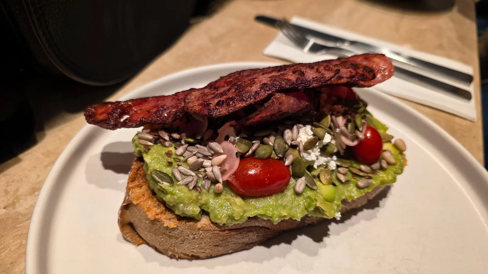 Smashed avocado on sourdough at Flow, scattered with seeds, feta, radish and cherry tomatoes, with turkey bacon laid across the top