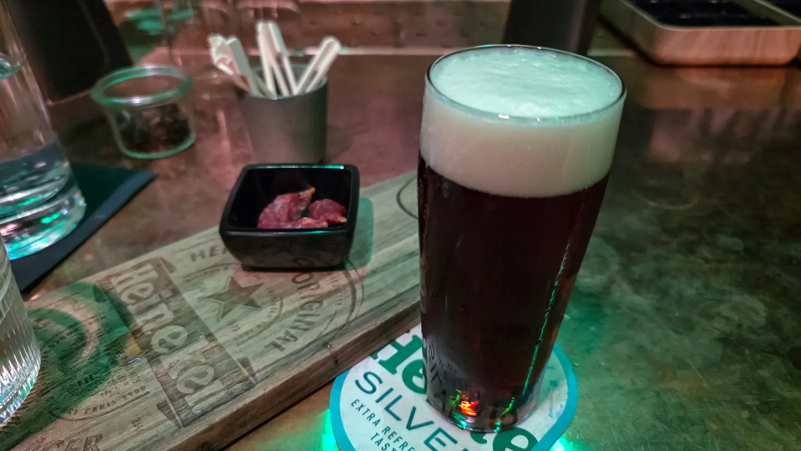 A dark red-brown beer under a thick head beside a small black dish of chorizo on a wooden board at Freddy’s bar