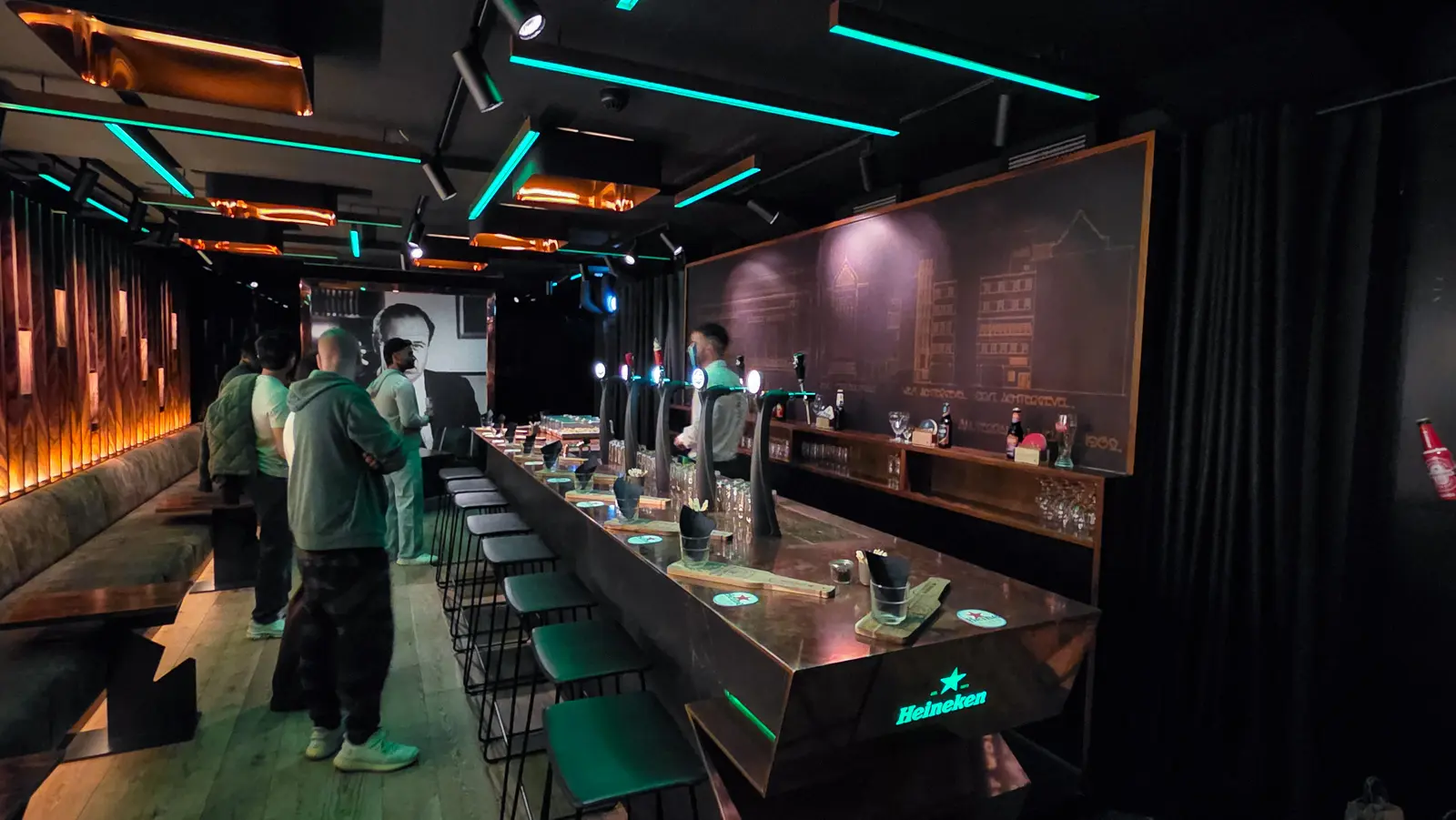 The long copper bar at Freddy’s under teal neon strips and copper ceiling panels, with black stools and guests standing at the far end