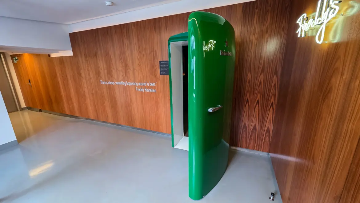 A green Heineken fridge standing open as a doorway in a walnut-panelled corridor, with a neon Freddy’s sign glowing on the wall