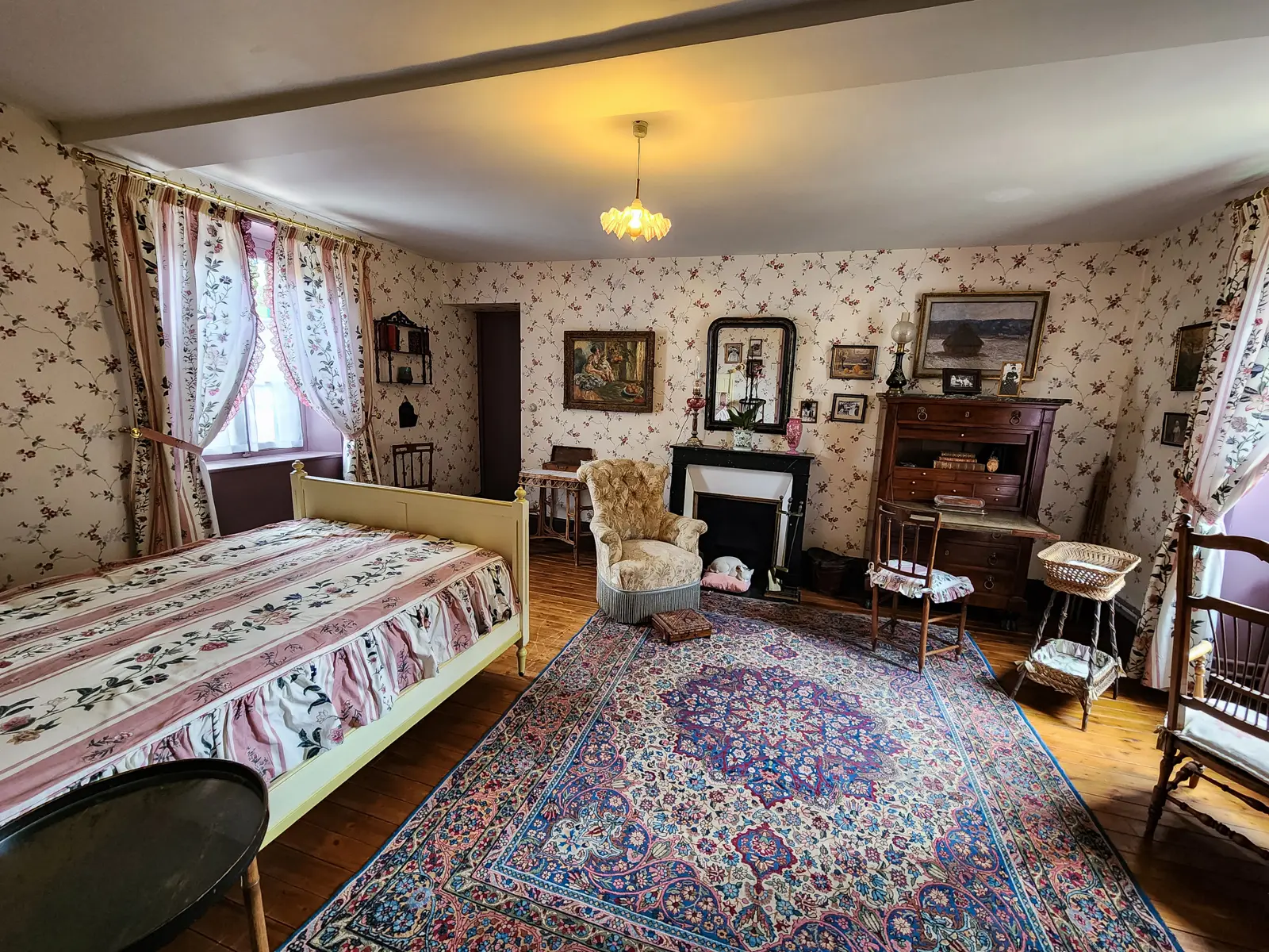 A bedroom at Monet’s house in Giverny, with floral wallpaper, a wooden bed, a marble fireplace and framed paintings on the walls