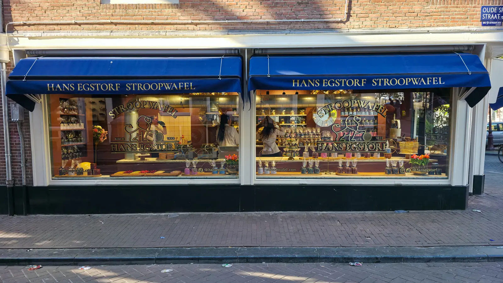 Two windows of Hans Egstorf under blue awnings on Oude Spiegelstraat in Amsterdam’s Nine Streets, with stacked stroopwafels and tins on display