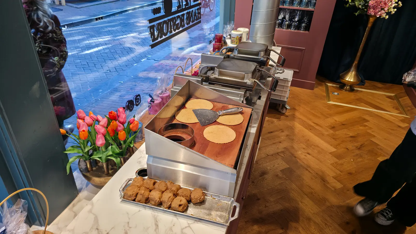 A waffle iron, a tray of dough balls and freshly pressed plain waffle rounds with a spatula at Hans Egstorf in Amsterdam, the street visible through the window behind
