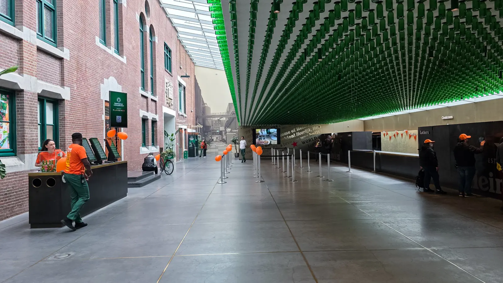 The arrival hall at the Heineken Experience, with a ceiling of green bottles above empty queue lanes and orange King’s Day balloons