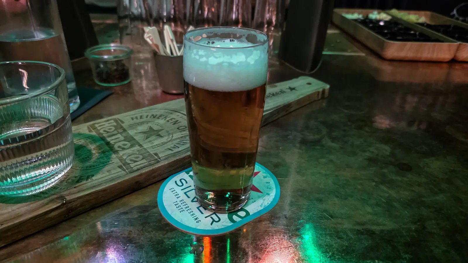 A glass of pale gold beer with a white head, standing on a coaster beside a branded wooden serving board at Freddy’s bar