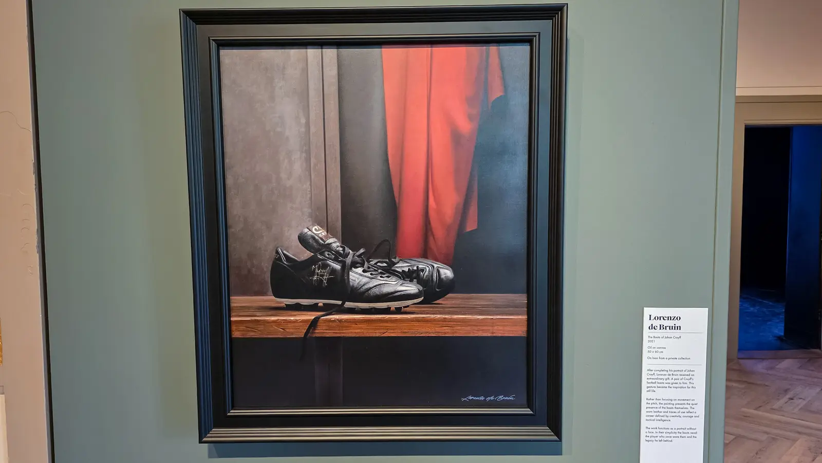 Lorenzo de Bruin’s painting The Boots of Johan Cruyff at the Moco Museum, a still life of worn black boots on a bench with a red shirt behind
