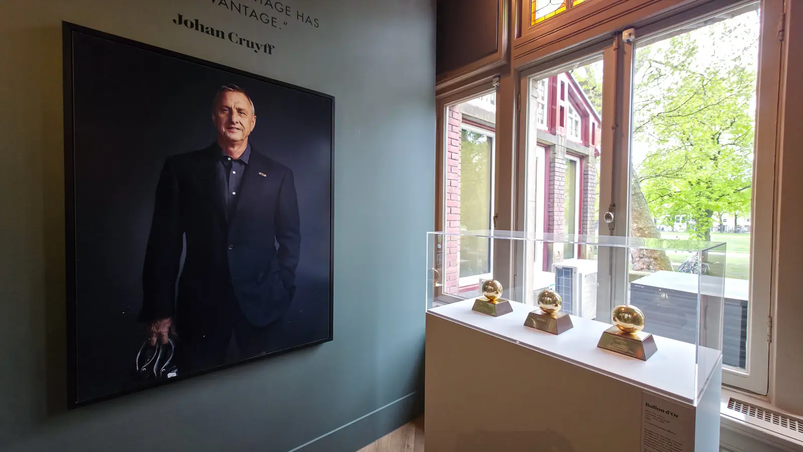 A large portrait photograph of Johan Cruyff in a dark suit at the Moco Museum, with a painted quotation above it and trophies in a case beside