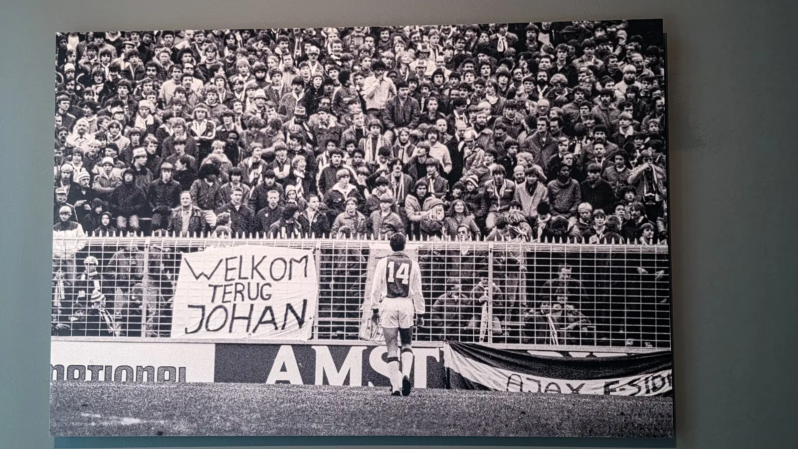 A large black and white photograph of Johan Cruyff in a number 14 shirt walking towards a crowd behind a fence, at the Moco Museum