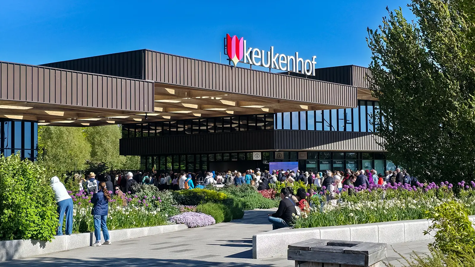 Visitors queue beneath the timber and dark-clad entrance building at Keukenhof, with ticket windows and flower beds in the foreground