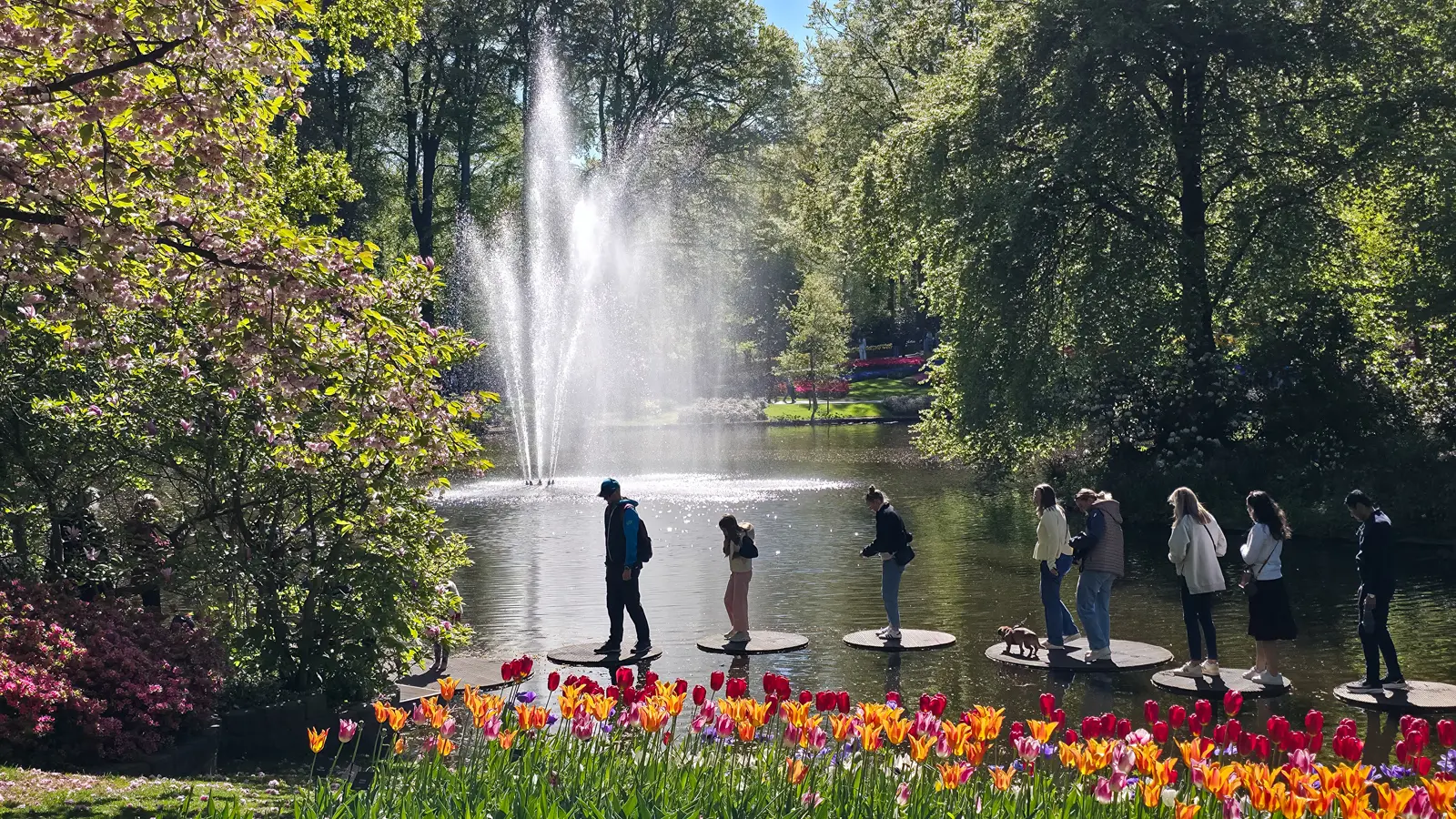 Visitors crossing circular stepping discs over the lake at Keukenhof, with a tall fountain behind and red and orange tulips in front