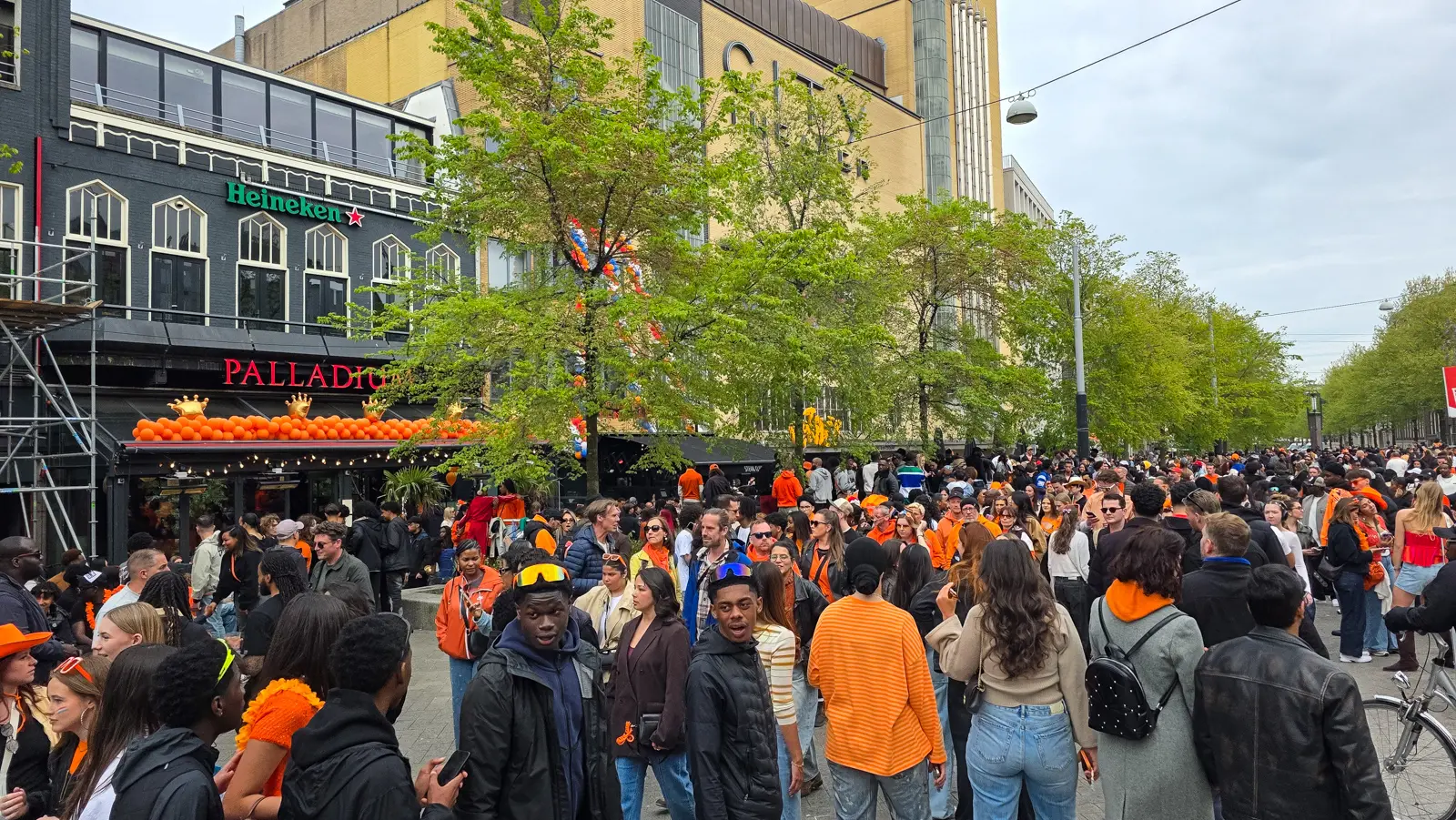 An arch of orange balloons topped with gold crowns above a bar entrance on Leidseplein, Amsterdam, a dense crowd in front and a Heineken sign above