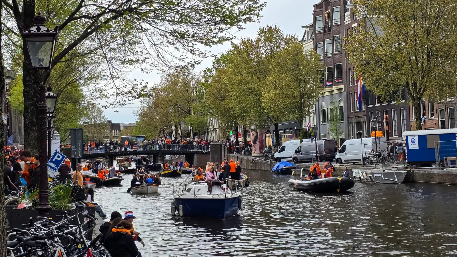 Boats full of people in orange filling an Amsterdam canal on King’s Day, with a crowded bridge behind and an inflatable dinghy alongside