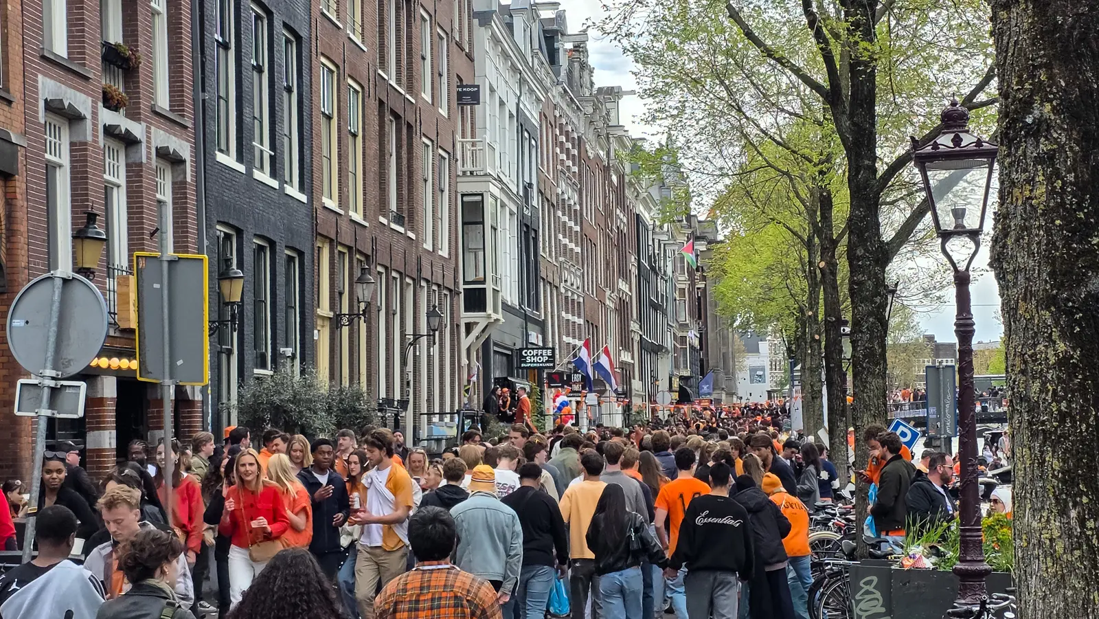 A crowded canal-side street in Amsterdam on King’s Day, Dutch flags on a lamppost, bars along the quay and a dense crowd in orange