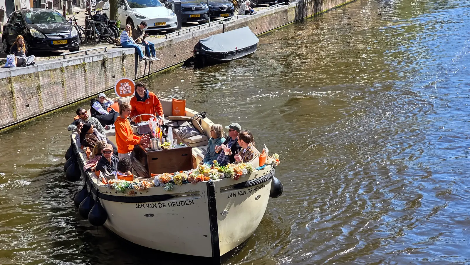 A decorated open canal cruise boat named Jan van de Heijden in Amsterdam, flowers along the bow, a bar set up on board and a guide steering