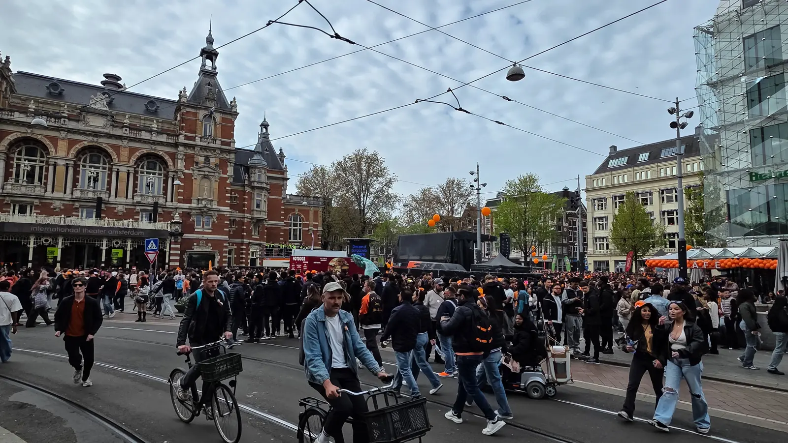 Crowds crossing the tram tracks on Leidseplein in Amsterdam on King’s Day, the red-brick Internationaal Theater Amsterdam behind under overhead tram wires
