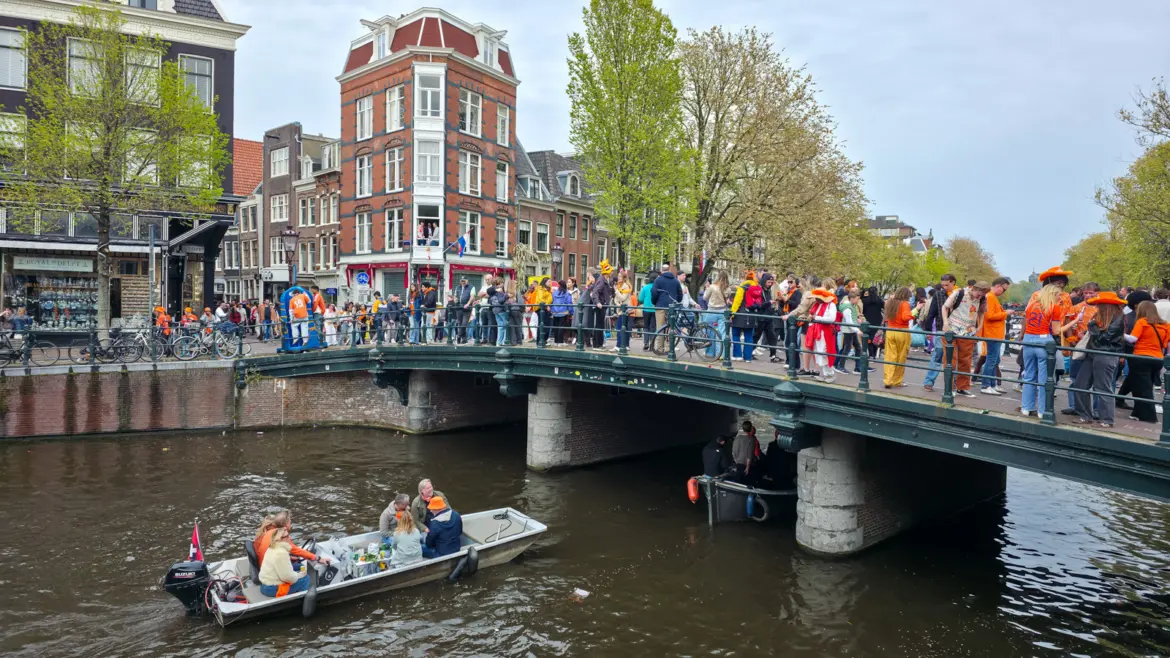 A canal bridge in Amsterdam packed with people in orange on King’s Day, a small open boat passing underneath