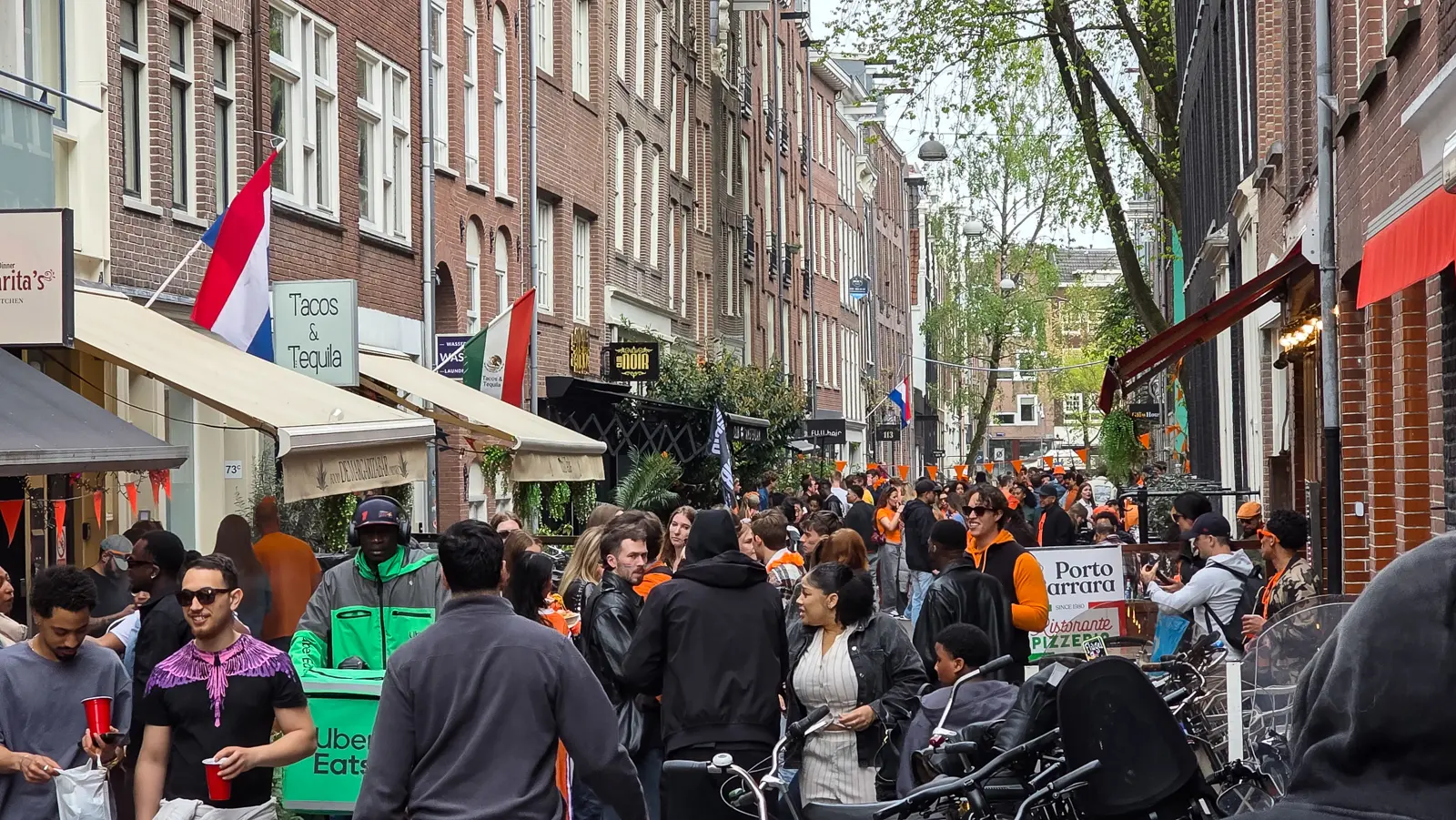 A narrow Amsterdam street crowded with people on King’s Day, hung with orange bunting and Dutch flags, a taco bar and pizzeria along one side
