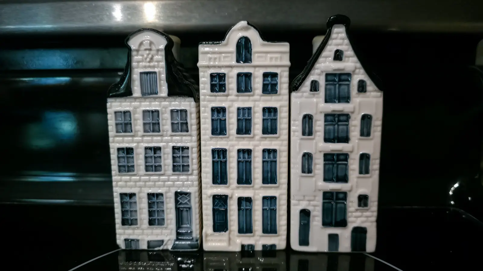 Three KLM Delft blue miniature houses in white and dark blue glaze, standing side by side on a dark reflective surface