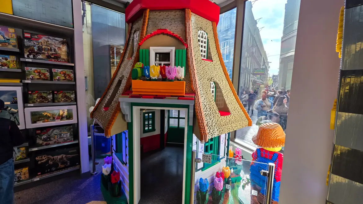 A brick-built Amsterdam canal house inside the LEGO Store, with a red roof, green shutters, a window box of brick tulips and a green parrot, its door open to the street